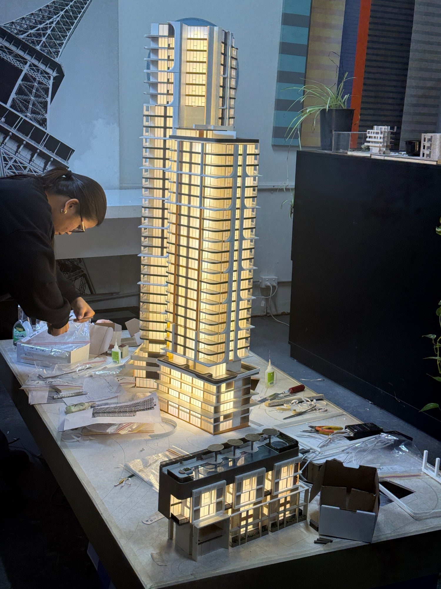 Person assembles illuminated apartment display model of a high-rise building on a cluttered studio table, architectural project.