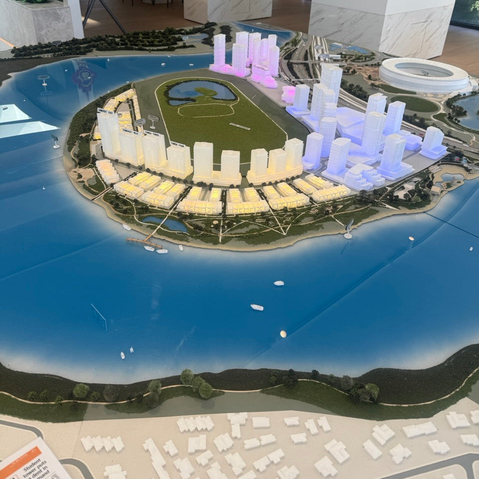 Architectural scale model of a waterfront development with illuminated buildings and landscaped green spaces on display.